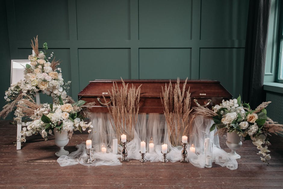 A somber floral arrangement displayed in front of a polished wooden casket, featuring cream and white roses, hydrangeas, and orchids accented with beige pampas grass. The flowers are arranged in elega
