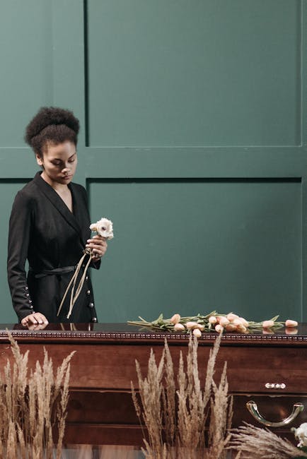 A young woman with curly dark hair and a serious expression, wearing a black blazer with button details, stands beside a dark wooden casket draped with a floral arrangement of light pink and cream ros