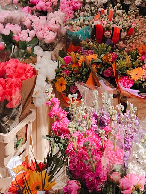 A vibrant display of fresh flowers, including pink and white roses, bright orange gerbera daisies, and clusters of pink stock, arranged in bouquets wrapped with colorful paper and transparent cellopha
