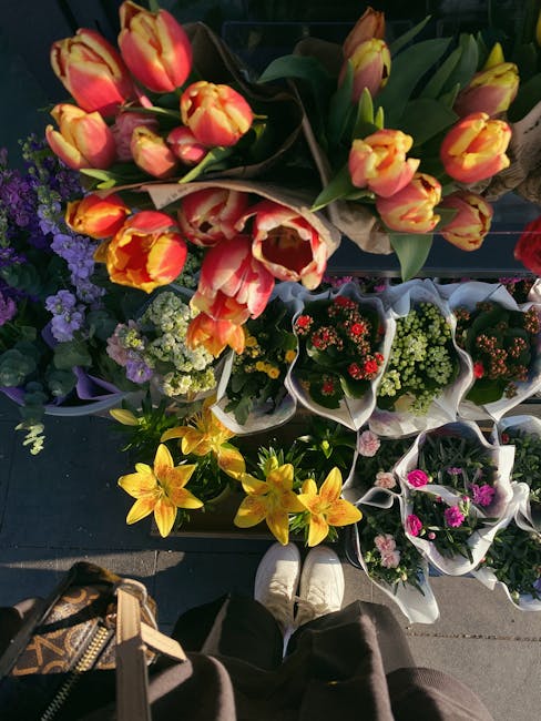 A top-down view of a variety of fresh floral bouquets arranged on a black surface, featuring a prominent bouquet of tulips with yellow and orange petals, green stems and leaves, wrapped in light brown