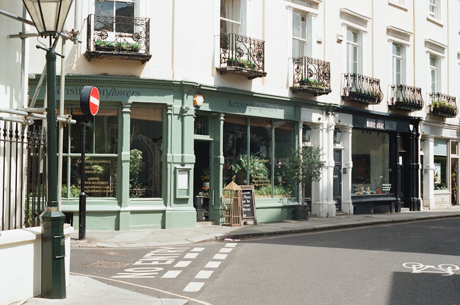 A street scene featuring a flower shop with a pale green facade and large glass windows displaying floral arrangements. The shop, named 'Kensington Flowers,' is situated on a corner with black wrought