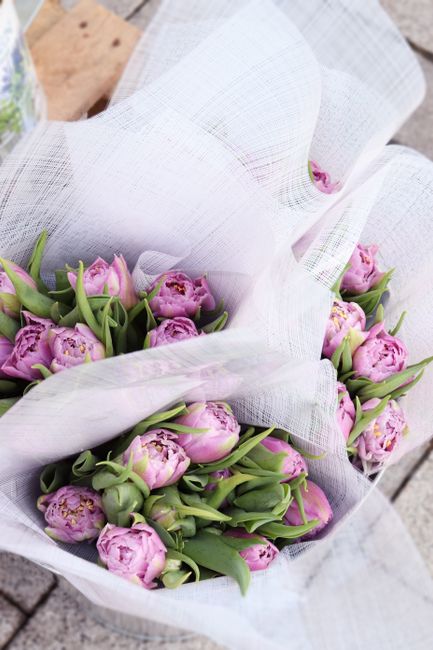 A bouquet of fresh pink tulips and peonies with green stems and leaves, arranged in a loose, natural style, wrapped in white textured tissue paper. The bouquet is resting on a rustic wooden surface, w