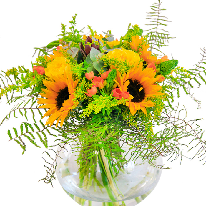 The first impression of this autumnal bouquet is one of sunlit exuberance, each element chosen with the care and keen eye of a local Clerkenwell florist. Sunflowers dominate, their golden yellow petals radiating outward in generous layers, reminiscent of Bloomsbury's golden afternoons. Nestled closely, classic yellow garden roses unwind their dense spirals, their warm hue evoking the buttery tones of pastries from Leather Lane's beloved bakeries. Between these blooms, glossy red hypericum berries twinkle, contrasted perfectly against the soft matte texture of the surrounding foliage and reminiscent of local market produce. Fern fronds spill over and down, their feathery edges moving softly like the breeze in Spa Fields. Pale sprigs, likely goldenrod, contribute intricate tiny yellow flowers, while slender spikes of greenery and cool eucalyptus leaves add depth and a gentle, almost herbal scent. The entire arrangement is grounded in a clear, spherical glass vase, where stems sit artfully tangled beneath the serene surface of fresh water, echoing Clerkenwell's tranquil side streets. This bouquet feels alive with joyful optimism-a perfect gift for milestones celebrated along Clerkenwell Road, or as a centerpiece brightening workspaces and gatherings, blending natural beauty with local charm.